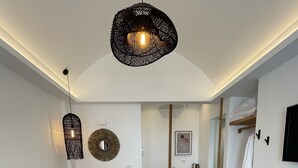 A modern bedroom with a large bed, a hanging light fixture, a round mirror, and a wooden bench. - Galatia Villas (Santorini)