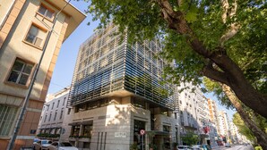 Un edificio de varios pisos con un diseño arquitectónico moderno, que combina fachada de vidrio y metal, y está rodeado de coches aparcados y árboles.