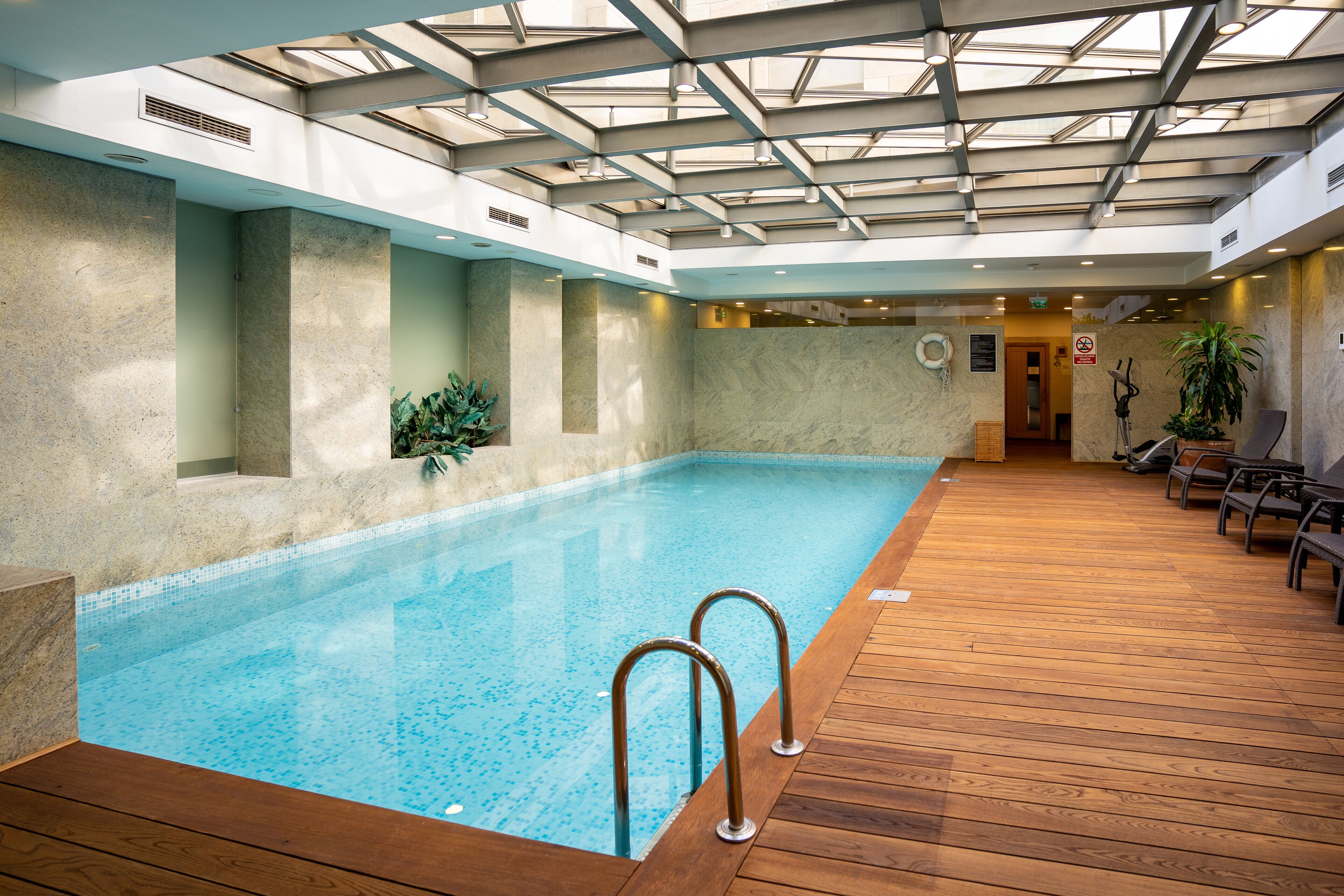 An indoor swimming pool with a wooden deck, a skylight ceiling, and a seating area with chairs.