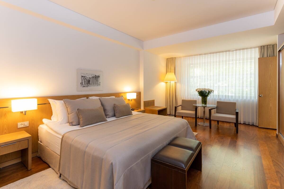 A hotel room with a large bed, bedside tables, a desk with a chair, a window with curtains, and a wooden wardrobe.