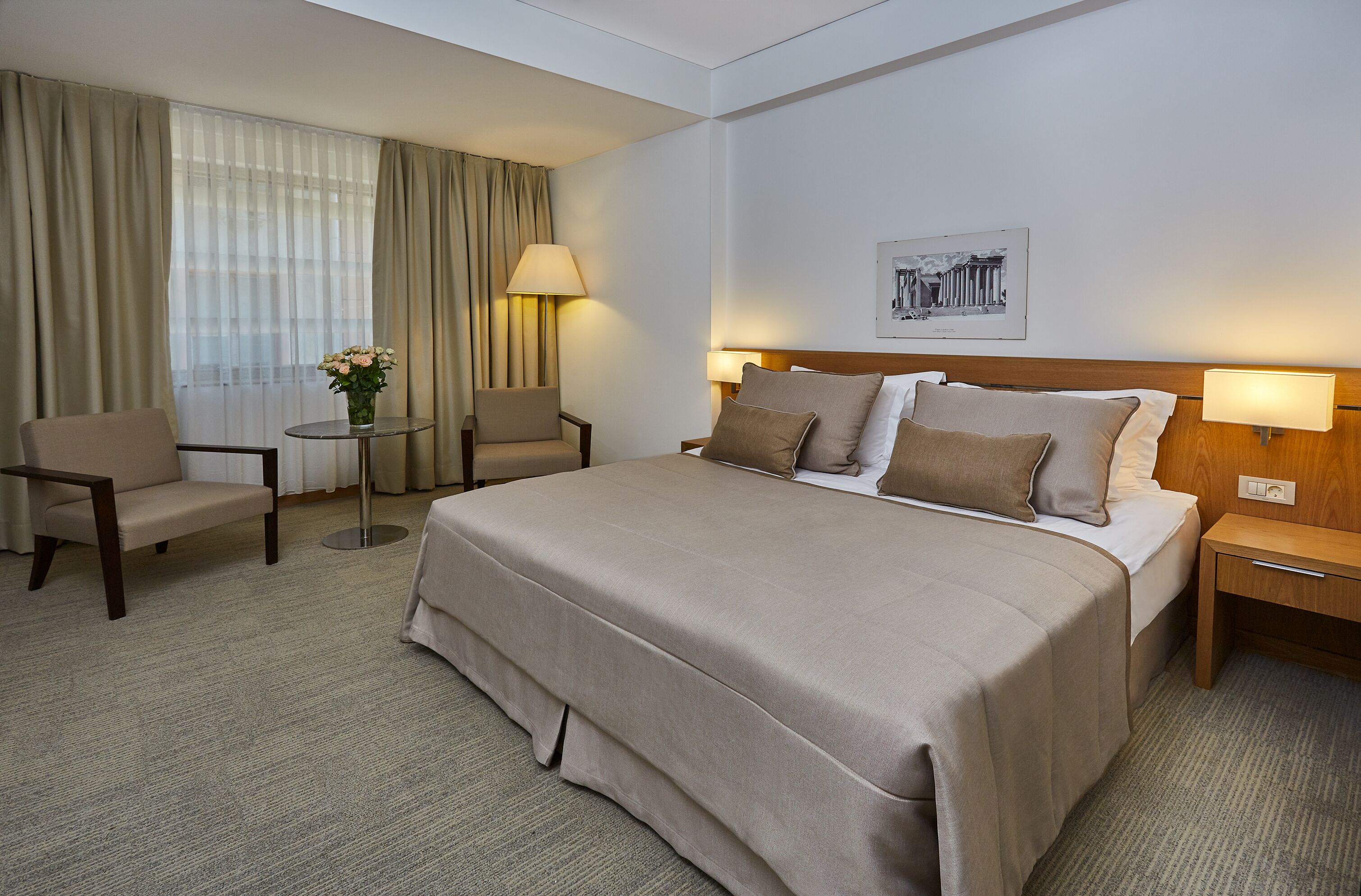 A hotel room with a large bed, two bedside tables, a chair, a small table with a lamp, and a window with curtains.