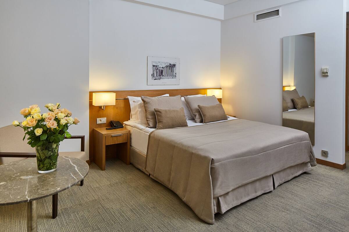 A hotel room with a bed, bedside lamps, a nightstand, a vase of flowers, a round table, and a wall-mounted picture.