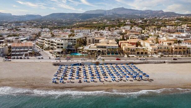 A coastal town with a beach featuring numerous blue umbrellas and a pool area. - Pearl Beach Hotel (Rethymno)