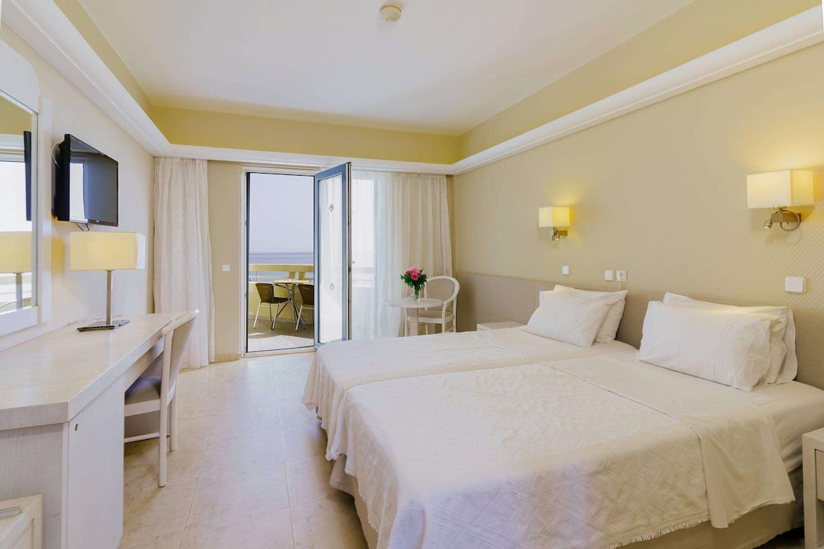 Habitación de hotel con dos camas, un escritorio, una silla, un televisor y un balcón con mesa y sillón.
