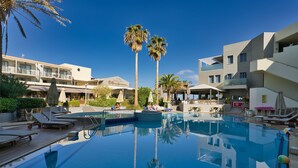 A swimming pool with lounge chairs and palm trees, surrounded by hotel buildings. - Pearl Beach Hotel (Rethymno)