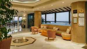 A spacious lobby with a large sofa, armchairs, and a coffee table. There are large windows offering a view of the sea. The lobby has a polished floor and a decorative rug. - Pearl Beach Hotel (Rethymno)