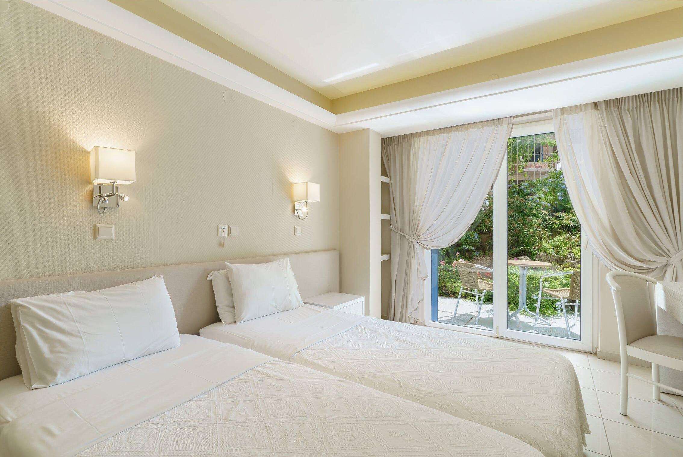 A hotel room with two beds, a balcony with a table and chairs, and a view of greenery outside.