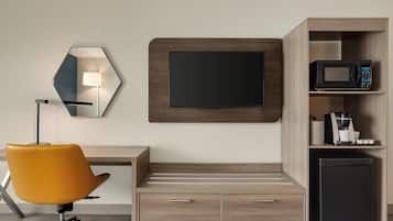 A modern hotel room with a wooden desk, a yellow office chair, a built-in microwave, a small refrigerator, a flat-screen TV, and a wall-mounted lamp.