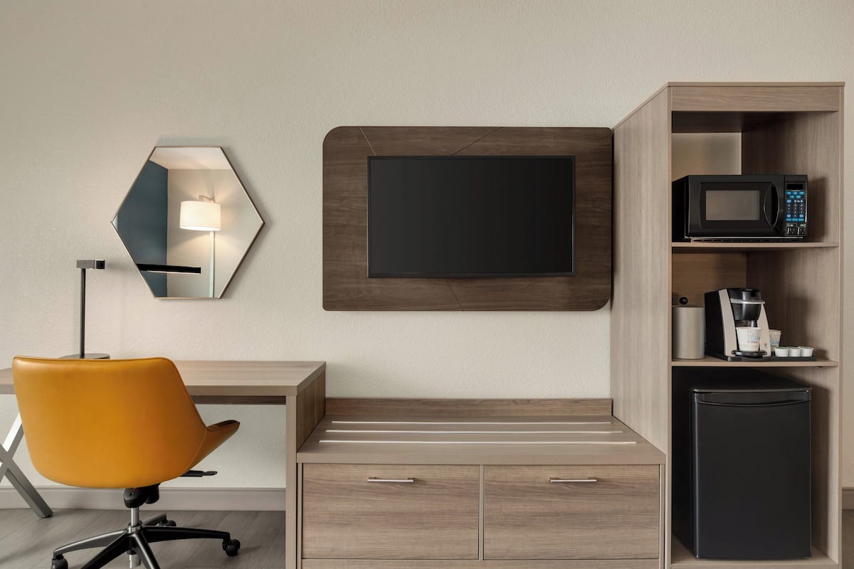 Une chambre d’hôtel moderne avec un bureau en bois, une chaise de bureau jaune, un four à micro-ondes encastré, un petit réfrigérateur, un téléviseur à écran plat et une lampe fixée au mur.