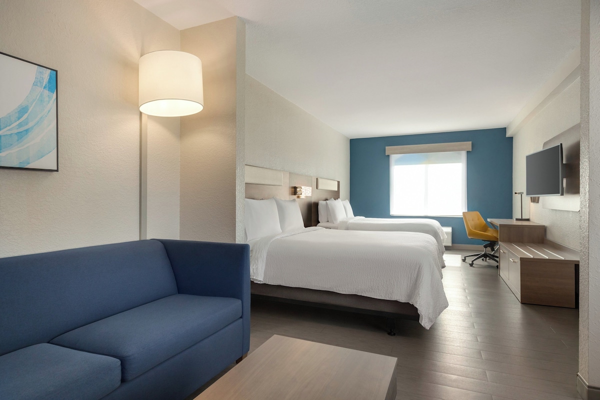 Une chambre d’hôtel avec un canapé bleu, deux lits avec du linge de lit blanc, un bureau avec une chaise et un téléviseur fixé au mur.