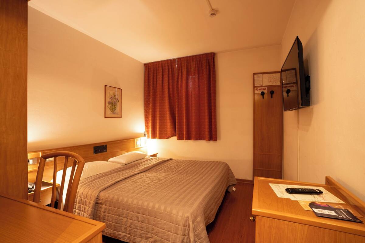A hotel room with a bed, a desk, a chair, a TV mounted on the wall, and a window with curtains.
