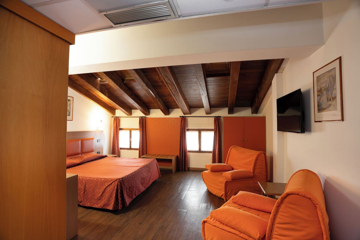 A hotel room with a wooden ceiling, a bed, two orange chairs, a small table, and a TV on the wall.