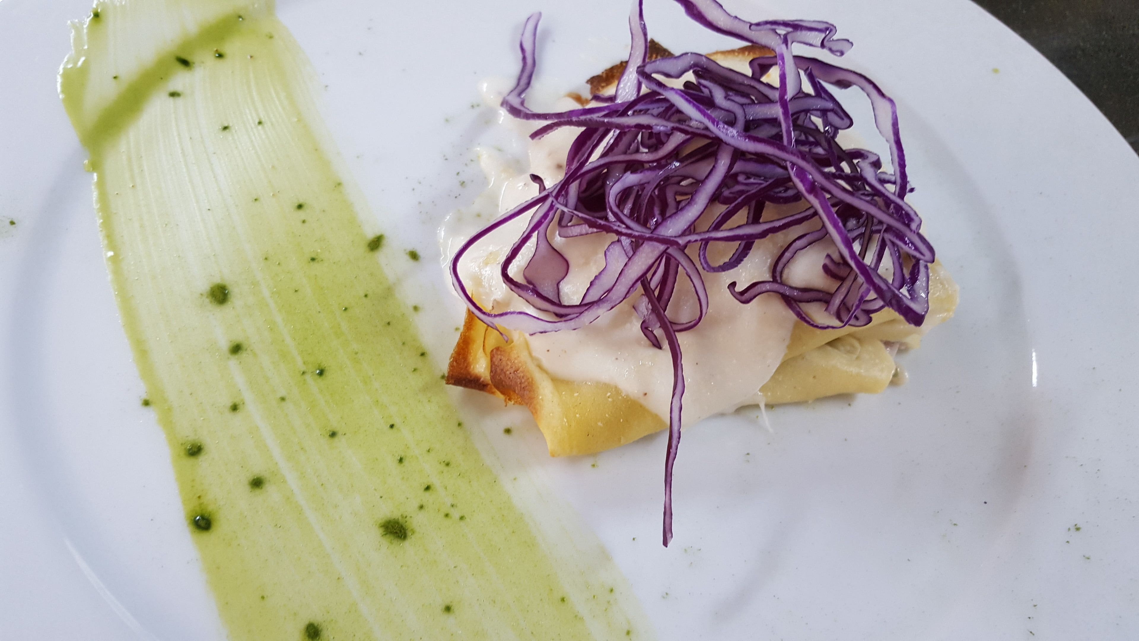 A plate with a piece of food topped with purple cabbage and a green sauce.