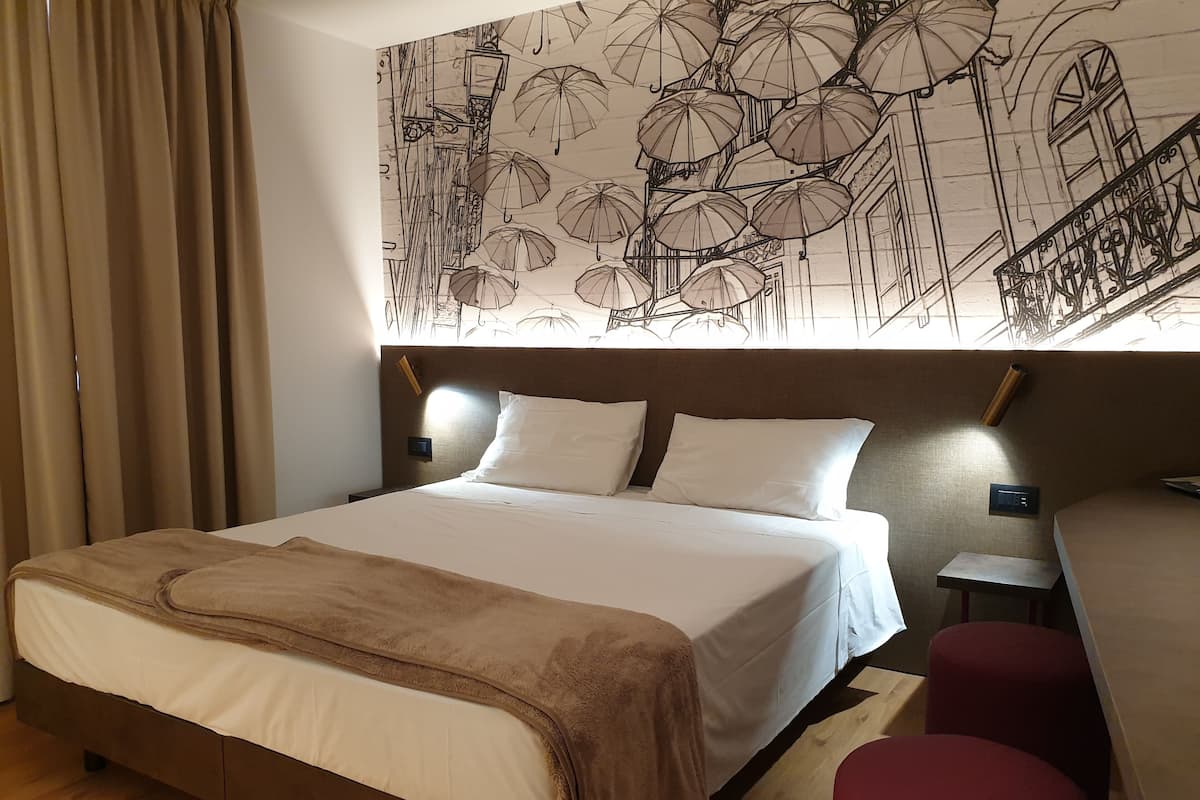 A hotel room with a large bed, a headboard with wall art, a bedside table with a lamp, and a small red chair.