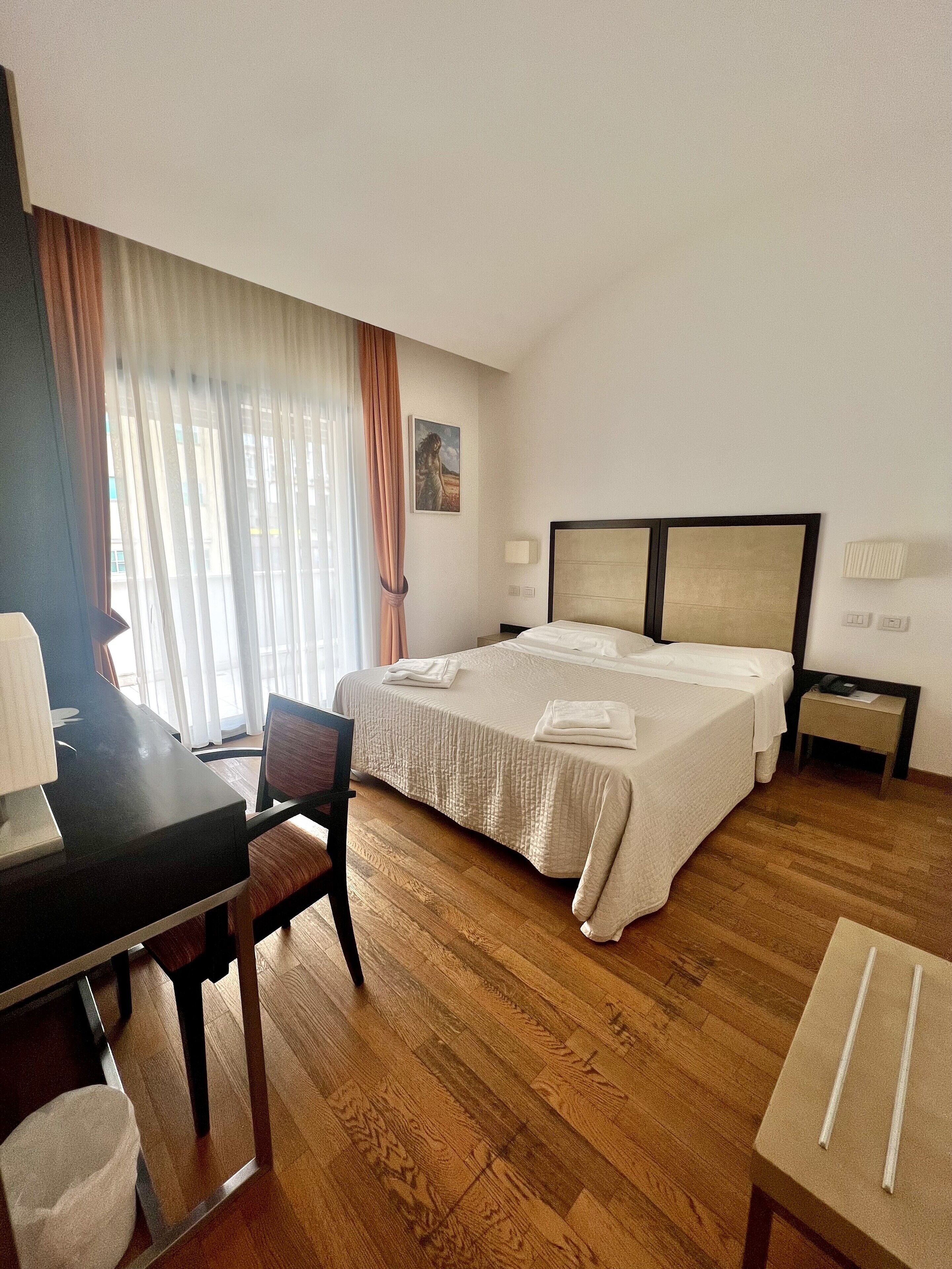 A hotel room with a bed, a desk, a chair, a nightstand, and a window with curtains.