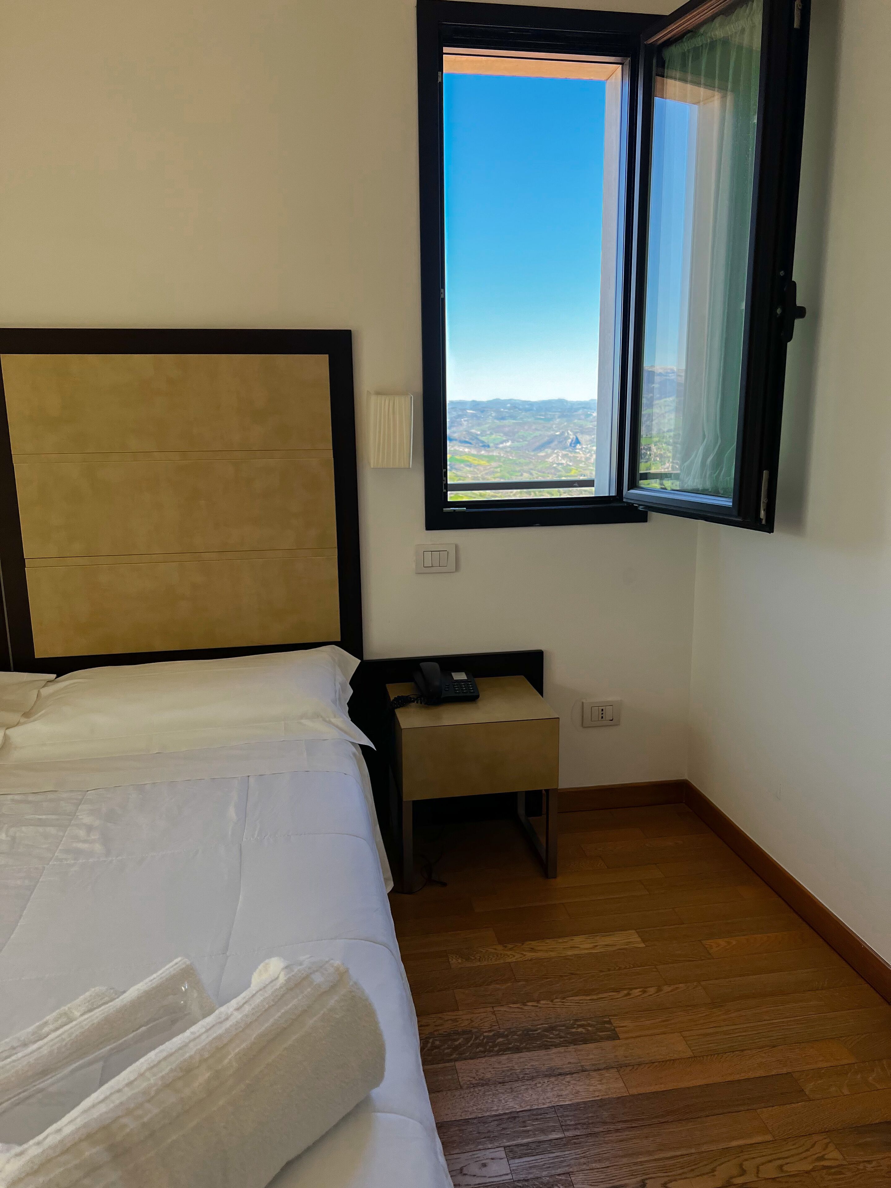 A hotel room with a bed, a bedside table with a phone, a window with a view of the landscape, and a wall-mounted light.