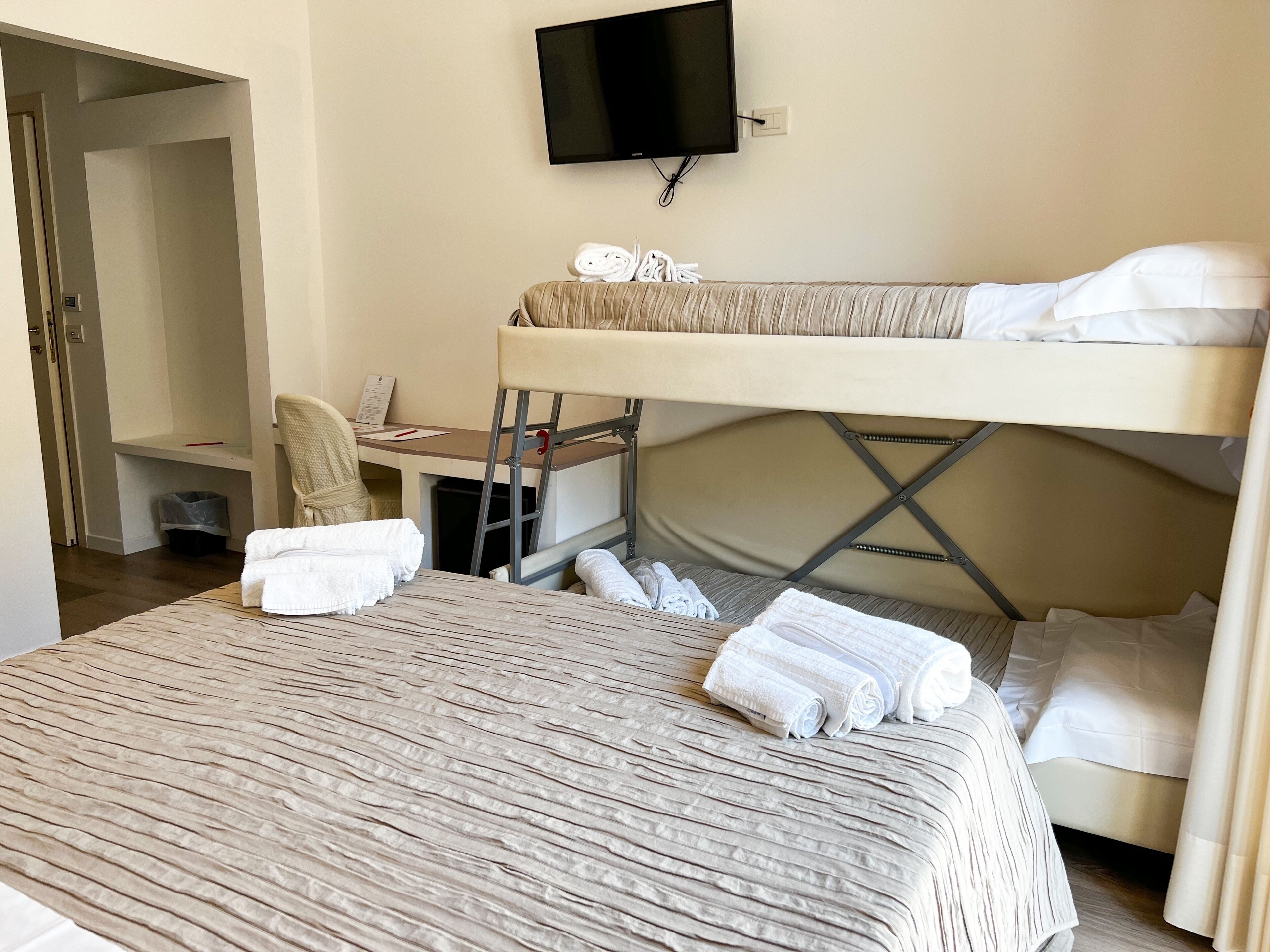 Una camera d'albergo con un letto a castello, una scrivania con una sedia, una televisione a parete e asciugamani piegati sul letto.
