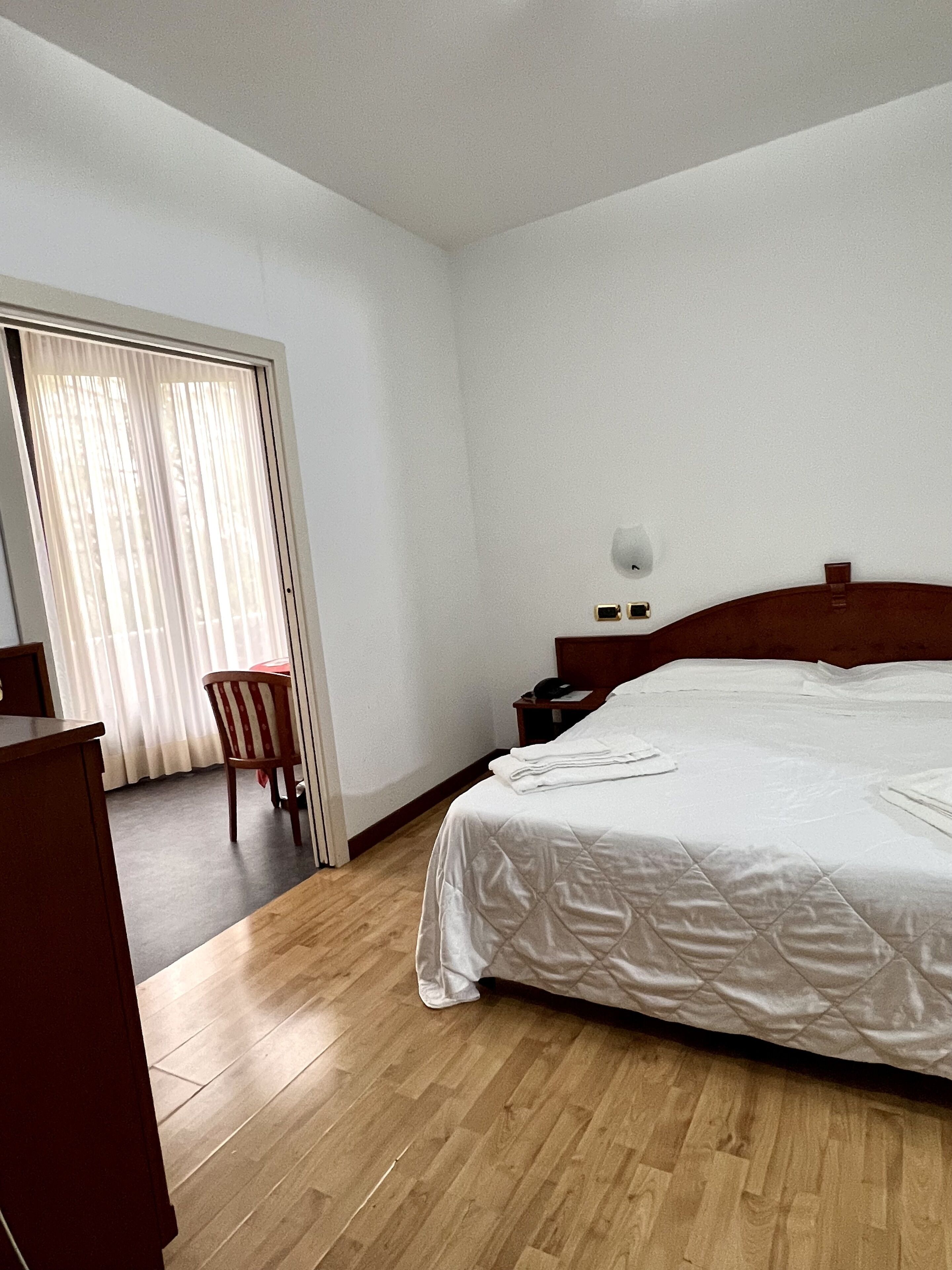 A hotel room with a wooden floor, a bed with white bedding, a wooden nightstand, a chair, and a window with curtains.