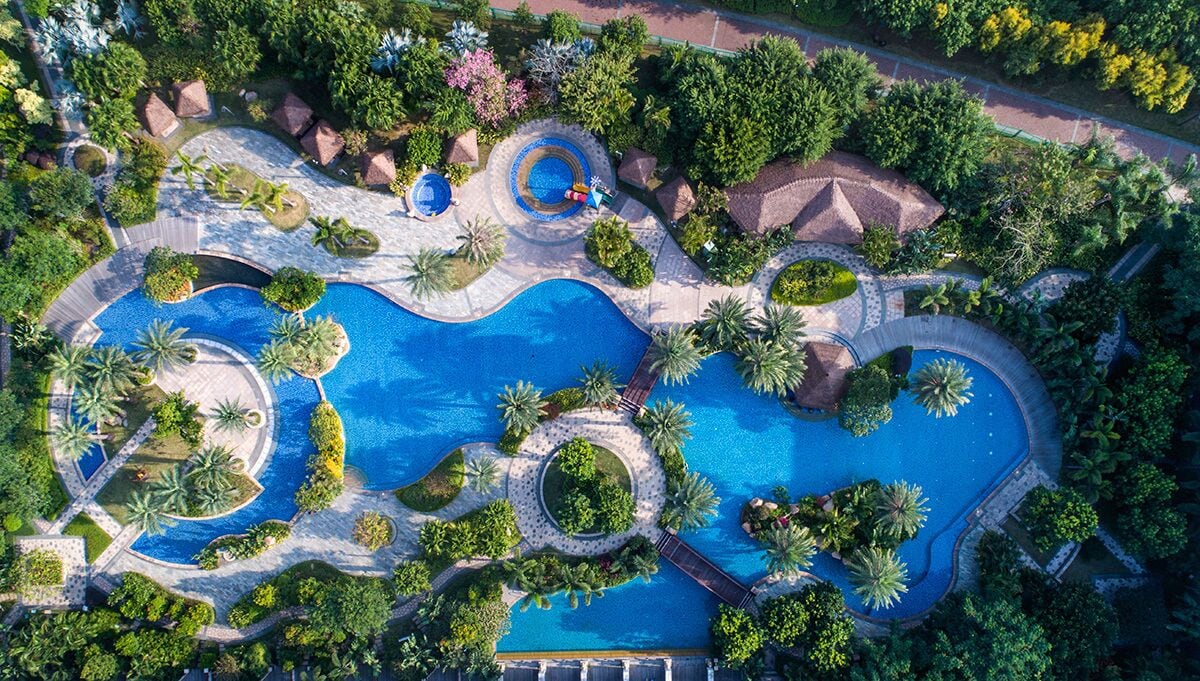 An aerial view of a resort with multiple swimming pools, surrounded by lush greenery and palm trees.