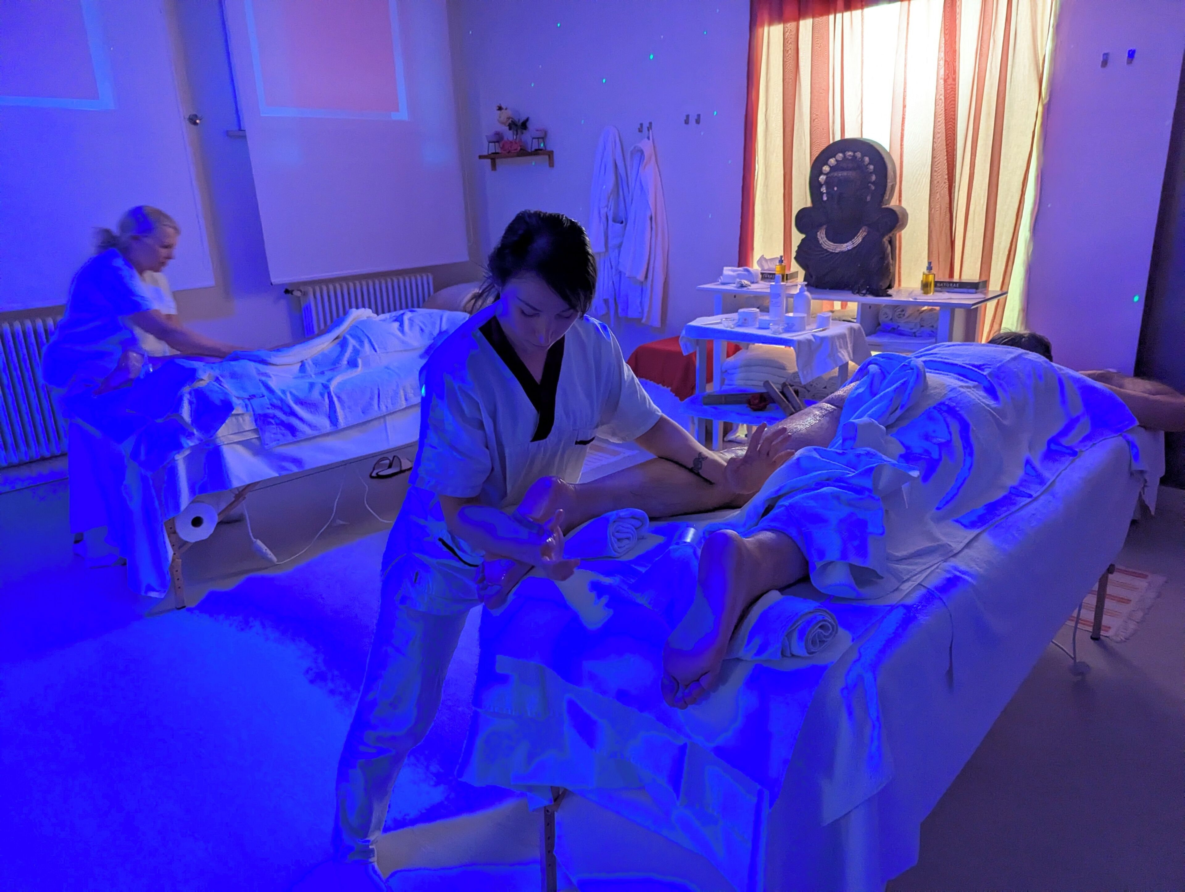 A room with a massage therapy bed, a massage therapist attending to a client, and another person in the background.