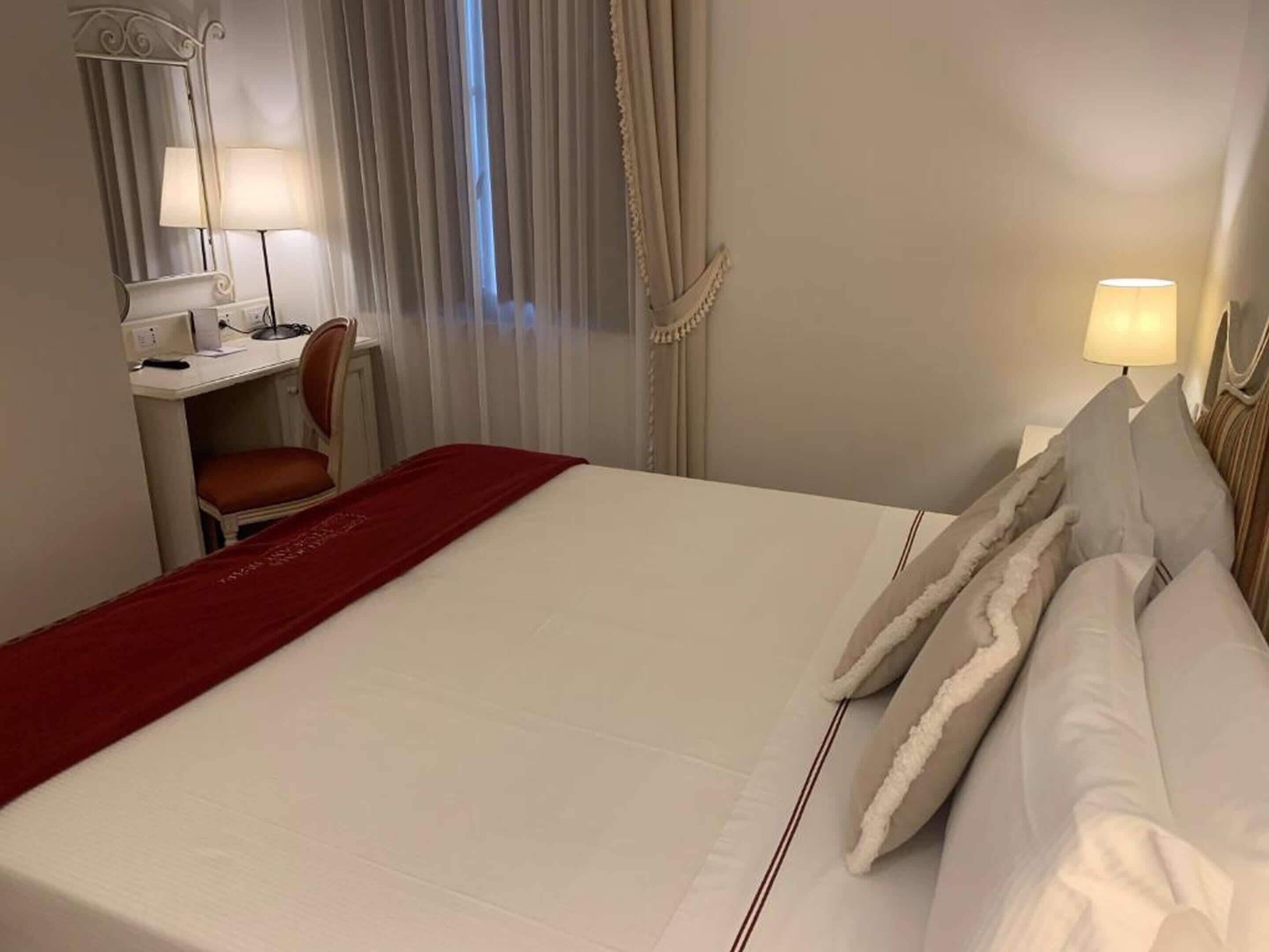 Una camera d'albergo con un letto grande, una scrivania con una sedia, una lampada e una finestra con tende.