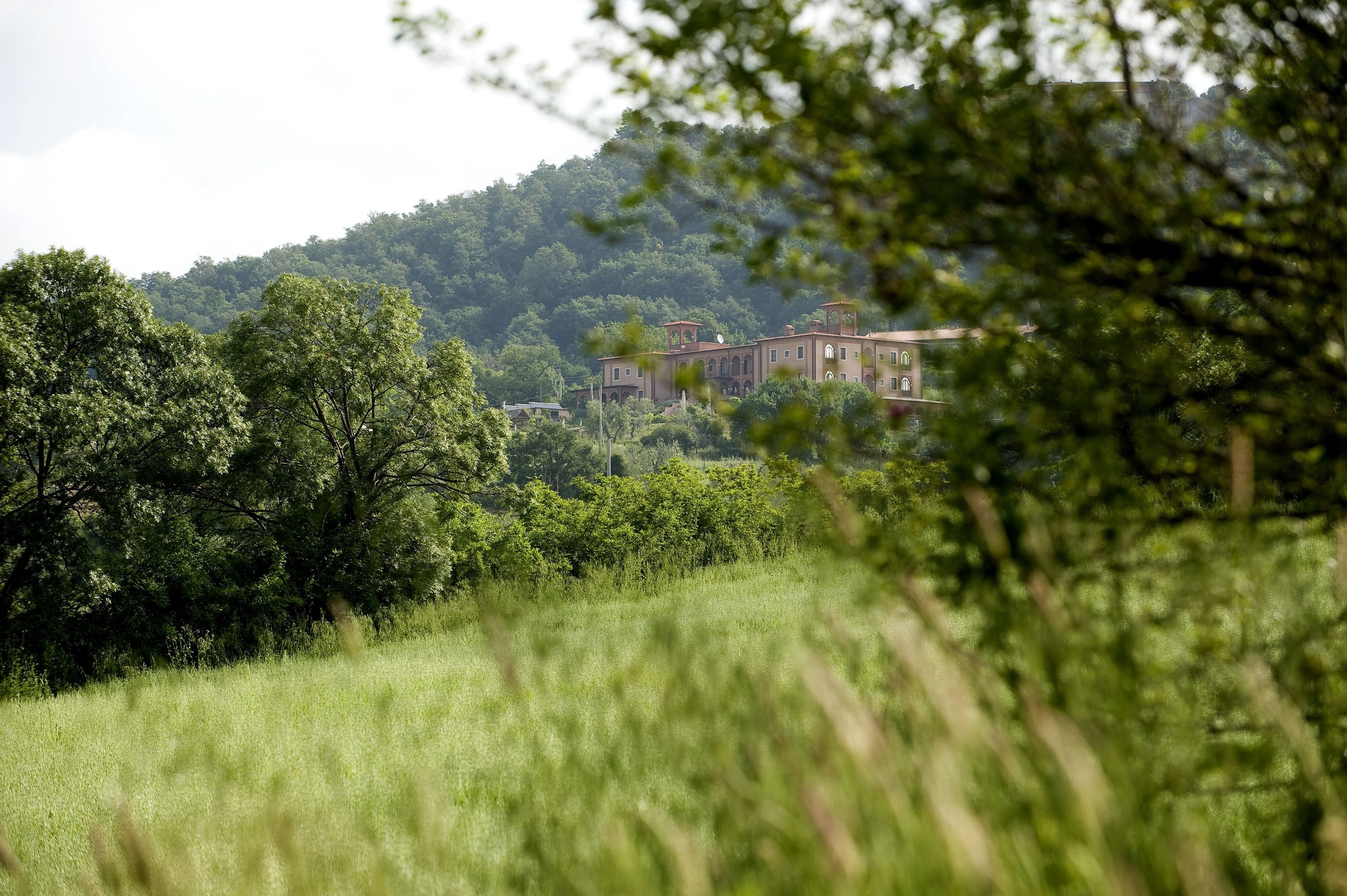 A building with a symmetrical facade, arched windows, and a central tower, surrounded by lush greenery and a hilly backdrop.