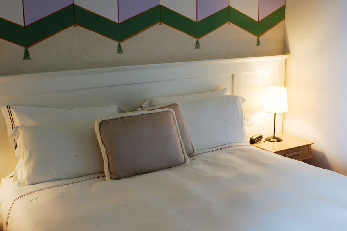 Un lit bien fait, avec du linge de lit blanc et deux oreillers, une lampe de chevet et un mur orné d’un motif géométrique.