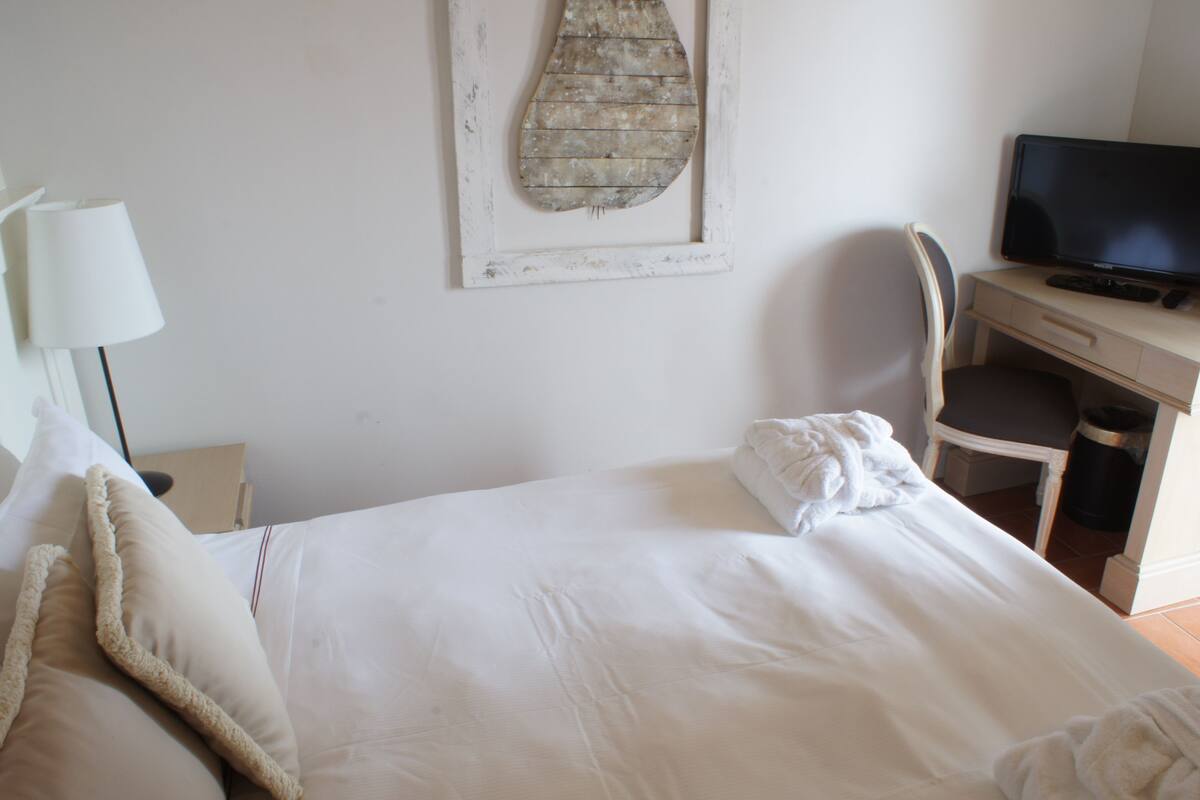Un lit bien fait, avec du linge de lit blanc et un oreiller moelleux, un bureau avec un ordinateur et une chaise, et une œuvre d’art accrochée au mur.