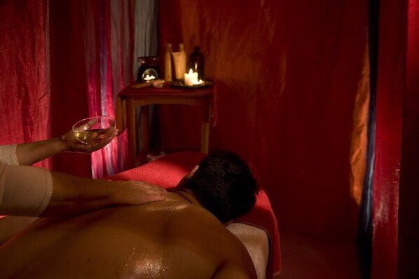 A person receiving a back massage in a room with red curtains and a small table with candles and bottles.