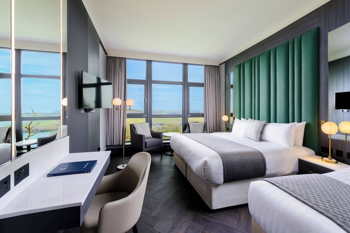 A modern hotel room with a large bed, a desk, and a chair. There is a TV mounted on the wall, and the room has large windows offering a view of the outdoors.