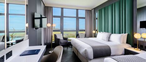 A modern hotel room with a large bed, a desk, and a chair. There is a TV mounted on the wall, and the room has large windows offering a view of the outdoors.