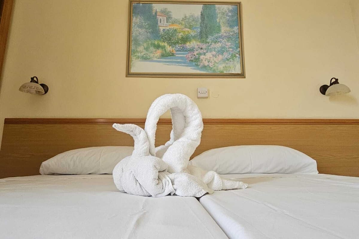 A bed with white linens, a painting of a garden on the wall, and two wall-mounted lamps.