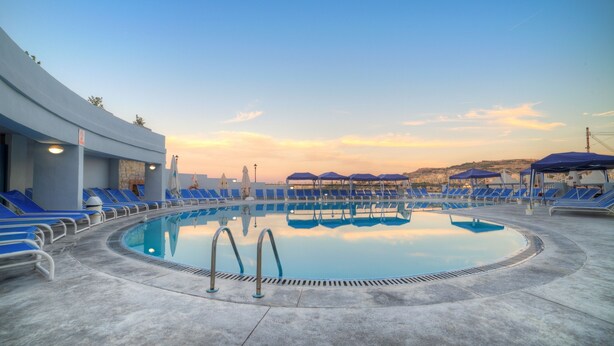 A pool area with blue lounge chairs and umbrellas, a clear sky, and a sunset. - Ambassador Hotel (St. Paul's Bay)