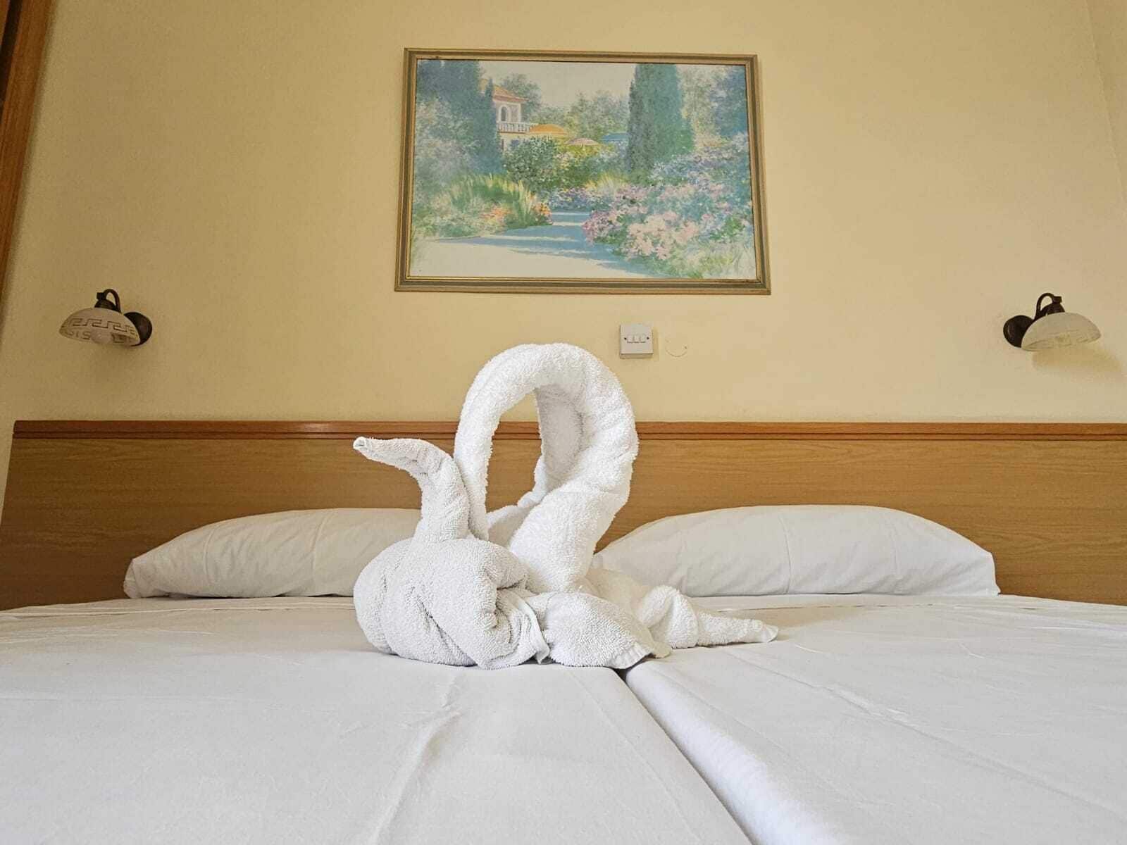 A bed with white linens, a painting of a garden on the wall, and two wall-mounted lamps.