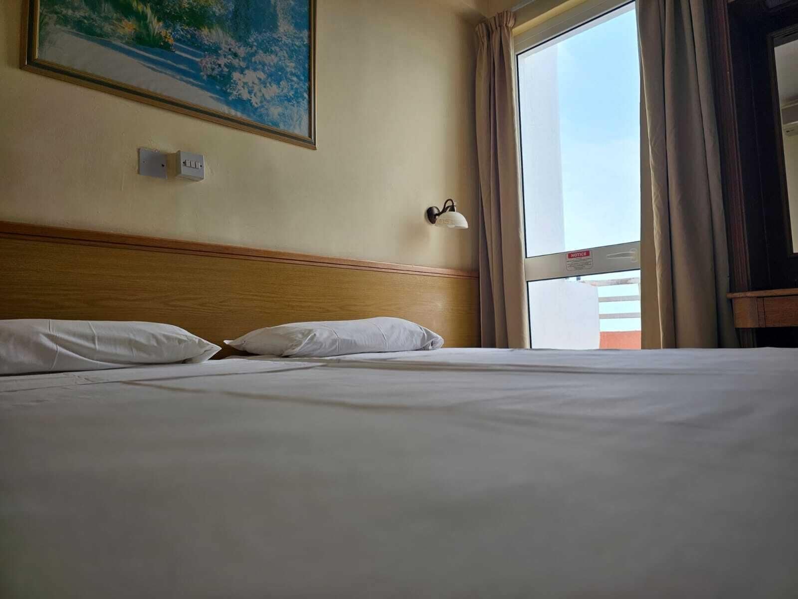 A hotel room with a bed, a painting on the wall, a window with curtains, and a notice sign.
