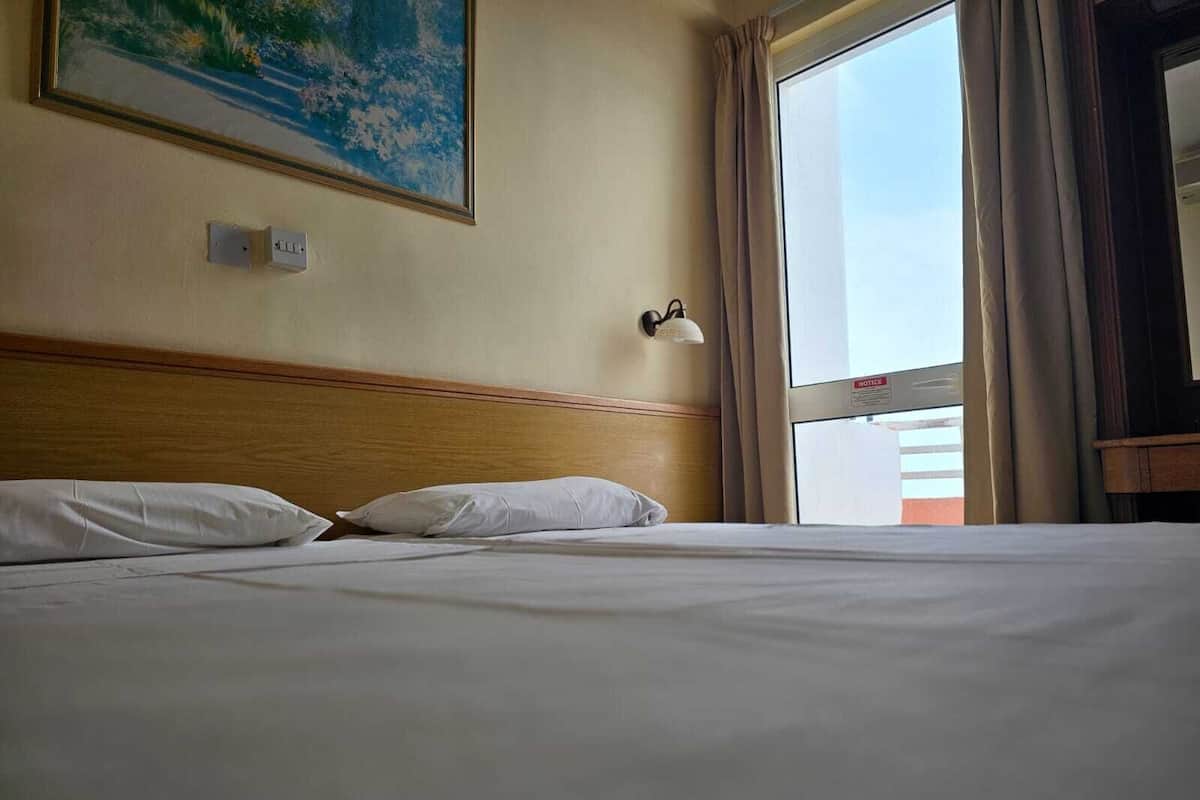 A hotel room with a bed, a painting on the wall, a window with curtains, and a notice sign.