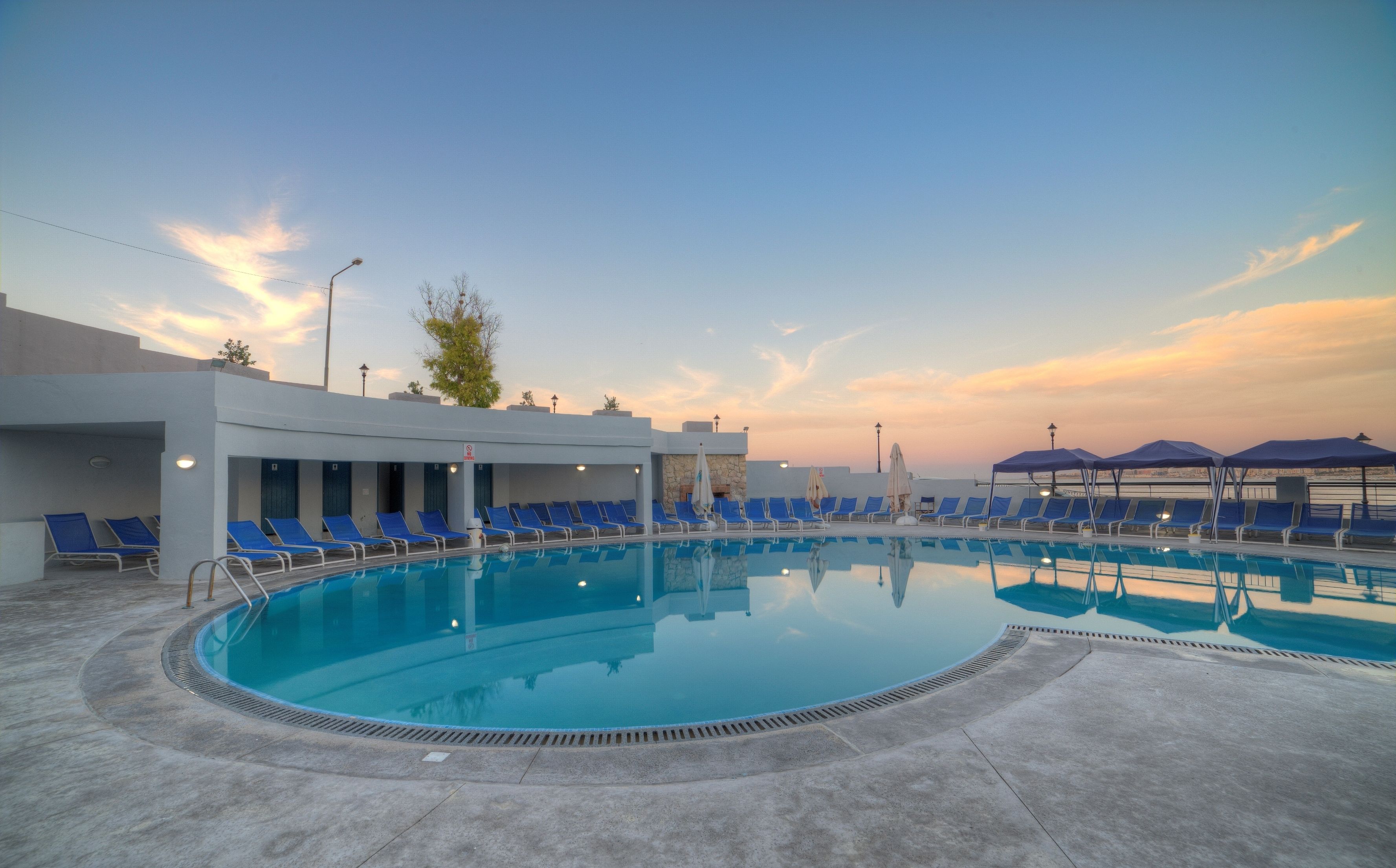 A swimming pool with blue lounge chairs and umbrellas, surrounded by a modern building with a stone facade.