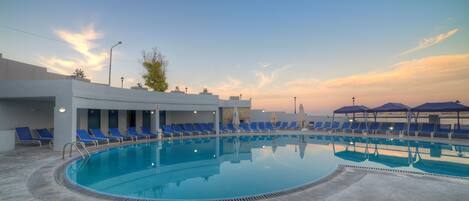 A swimming pool with blue lounge chairs and umbrellas, surrounded by a modern building with a stone facade.