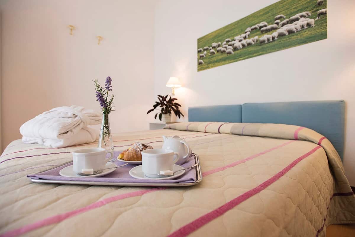 A neatly made bed with a tray of breakfast items, a vase with flowers, and a painting of sheep on the wall.