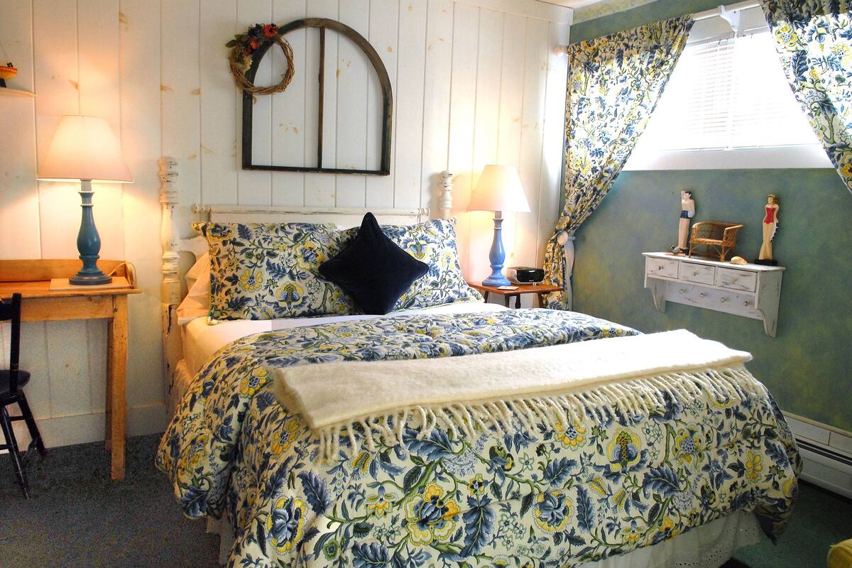 Une chambre à coucher avec une couvre-lit à motifs floraux, deux lampes de chevet, une table de chevet en bois, une chaise et une commode ornée de statuettes.
