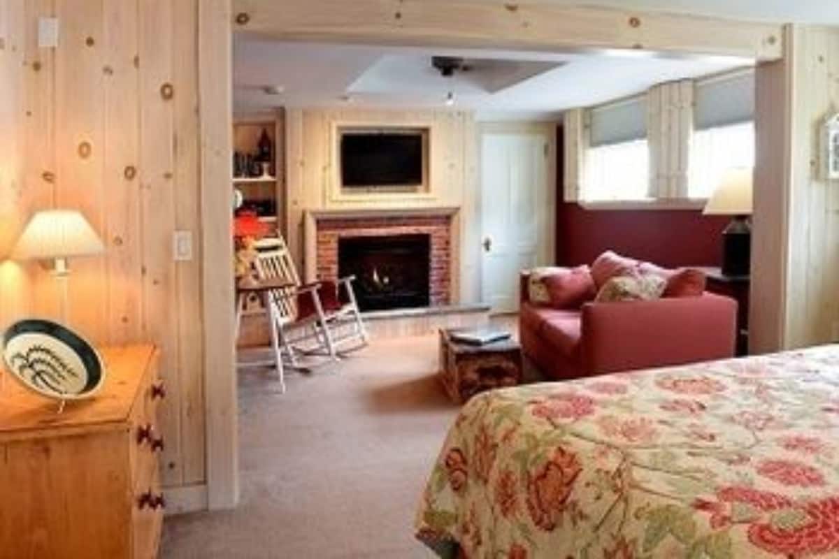 Une pièce chaleureuse avec une cheminée, un lit recouvert d’une couvre-lit à motifs floraux, un fauteuil rouge et une commode en bois.
