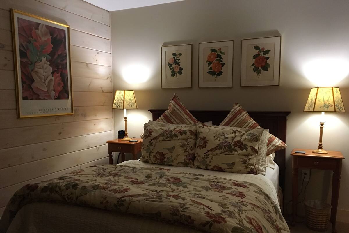 Une chambre à coucher avec une tête de lit en bois, une literie à motifs floraux, deux lampes de chevet et trois tableaux à motifs floraux encadrés accrochés au mur.