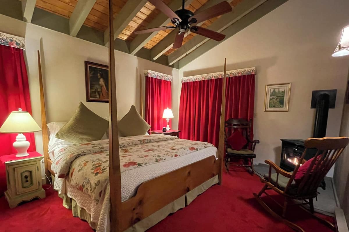 Un lit à baldaquin avec des rideaux rouges, un plancher en bois, une cheminée et un fauteuil à bascule.