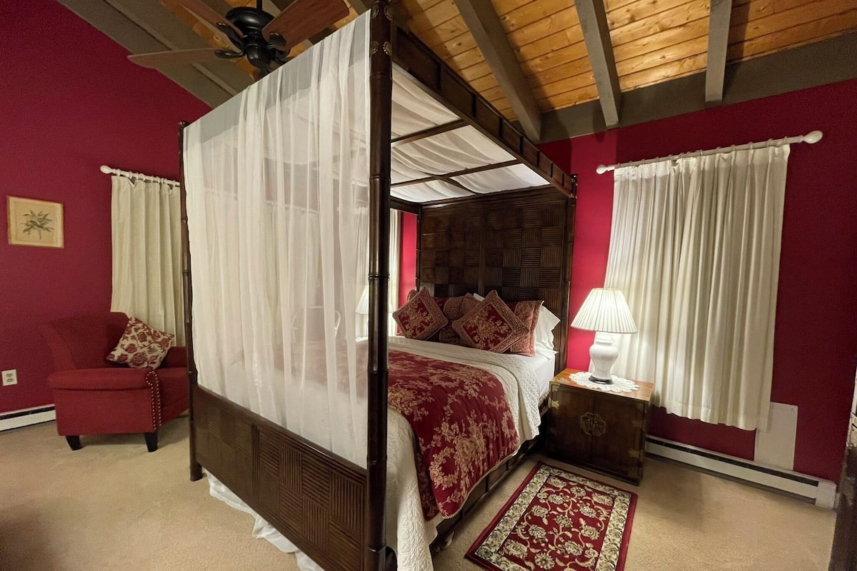 Un lit à baldaquin, un canapé rouge, une table de chevet avec une lampe et un tapis à motifs dans une pièce aux poutres apparentes et aux murs rouges.