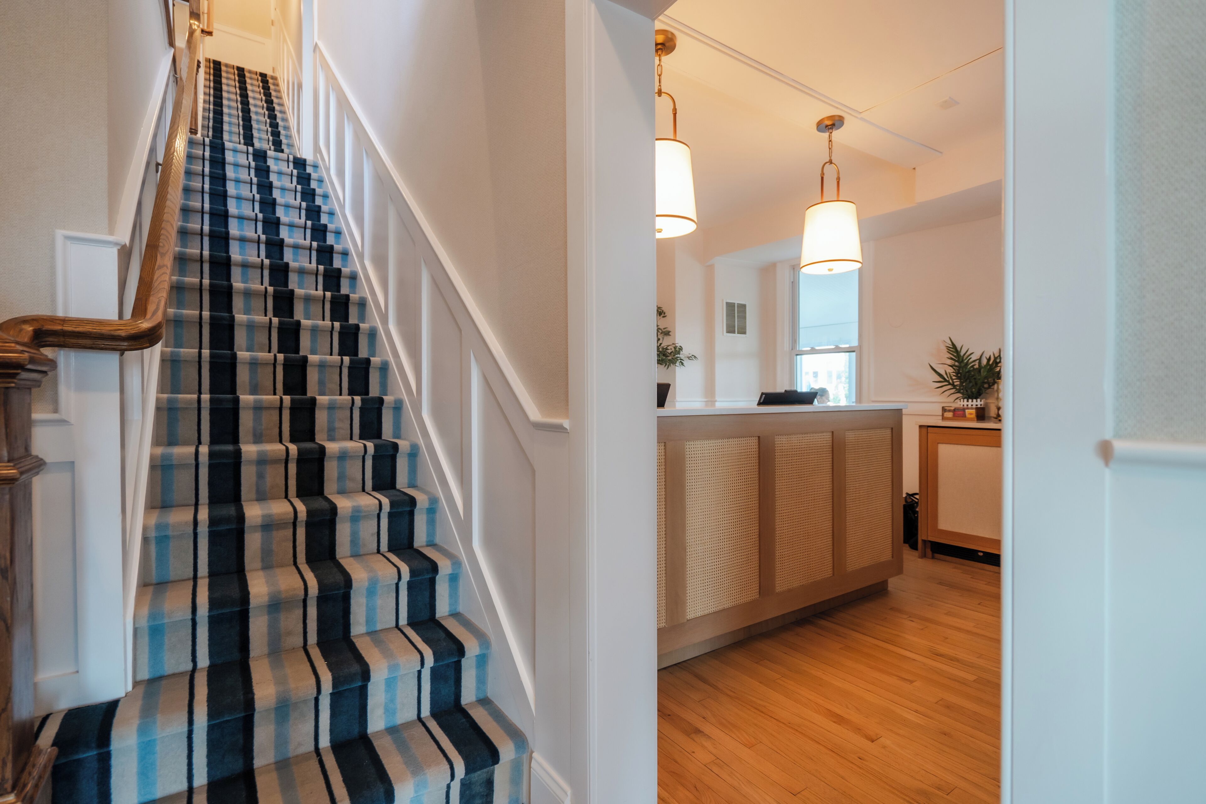 A staircase with a checkered carpet, a wooden handrail, and a kitchen area with a wooden floor and pendant lighting.