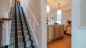 A staircase with a checkered carpet, a wooden handrail, and a kitchen area with a wooden floor and pendant lighting.