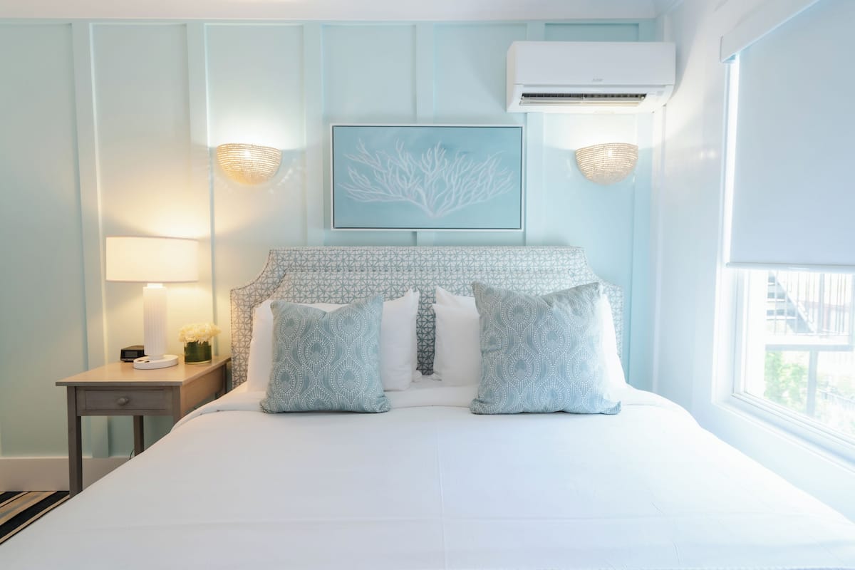 Ein Schlafzimmer mit einem Bett, einem Nachttisch, einer Lampe, einer Klimaanlage und einem gerahmten Kunstwerk an der Wand.