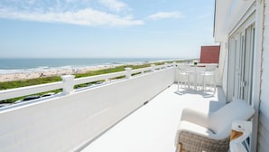 Un balcón con vistas a la playa, muebles de exterior blancos y una silla de mimbre.