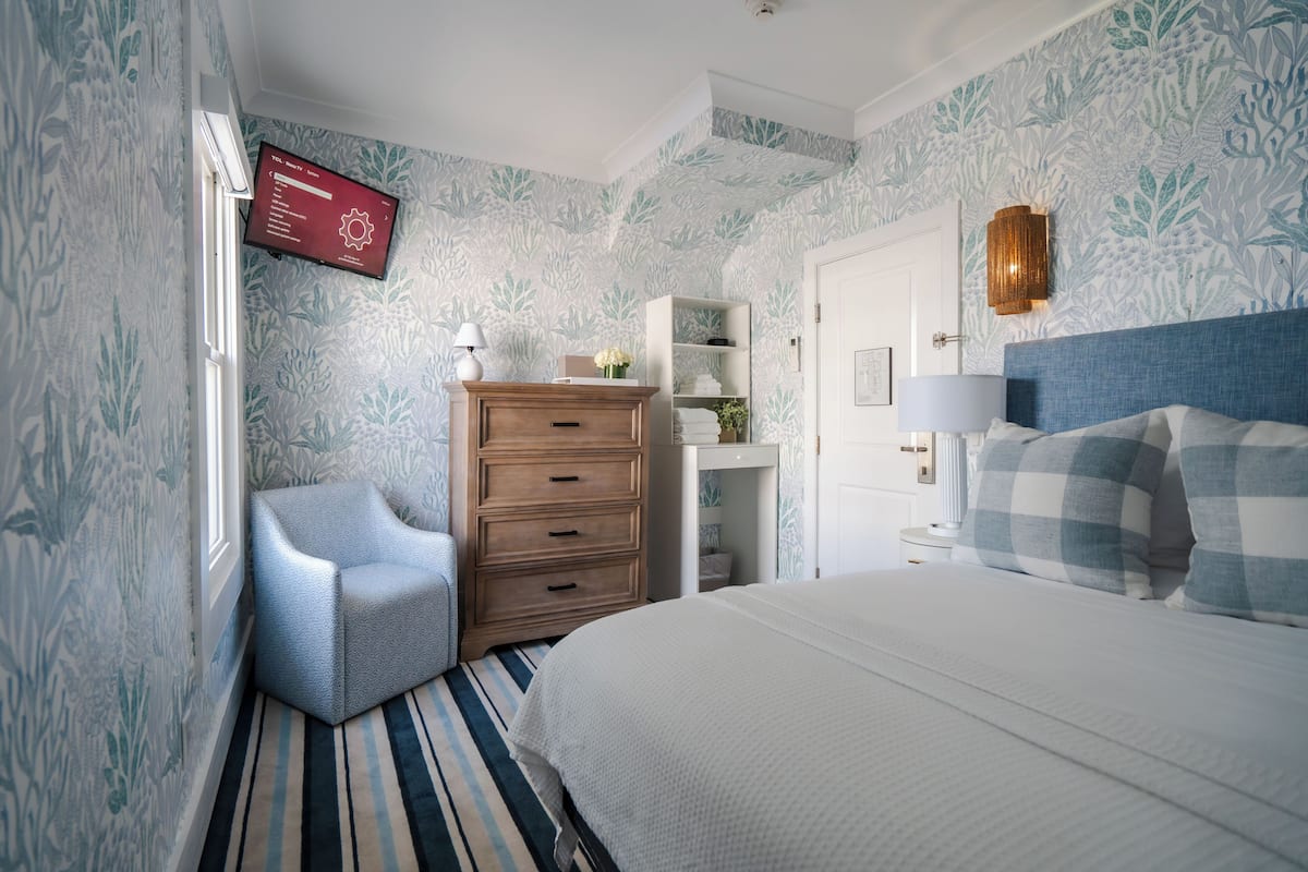 Ein Schlafzimmer mit einem Bett, einem Schminktisch, einem Sessel, einem an der Wand befestigten Fernseher und einer Tür mit Spiegel.