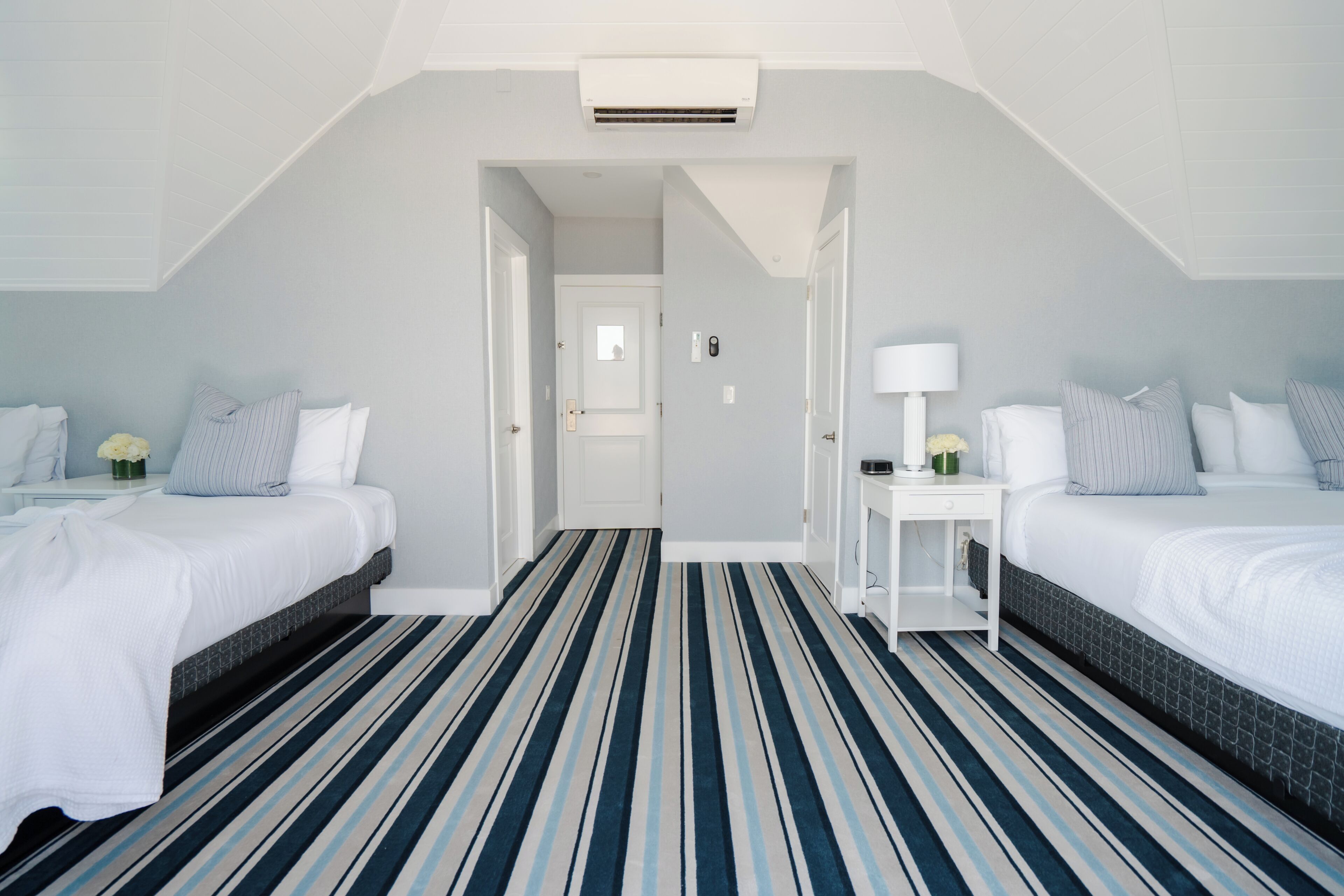 Ein Hotelzimmer mit zwei Betten, einem Streifen-Teppich, einer weißen Tür und einem kleinen Nachttisch mit einer Lampe.